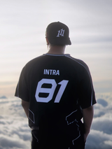 A man viewed from behind wearing a black athletic jersey with 'INTRA 01' printed in white on the back, along with a black baseball cap featuring a white monogram 111 angel number logo. He is standing outdoors above a sea of clouds, looking out over the horizon.