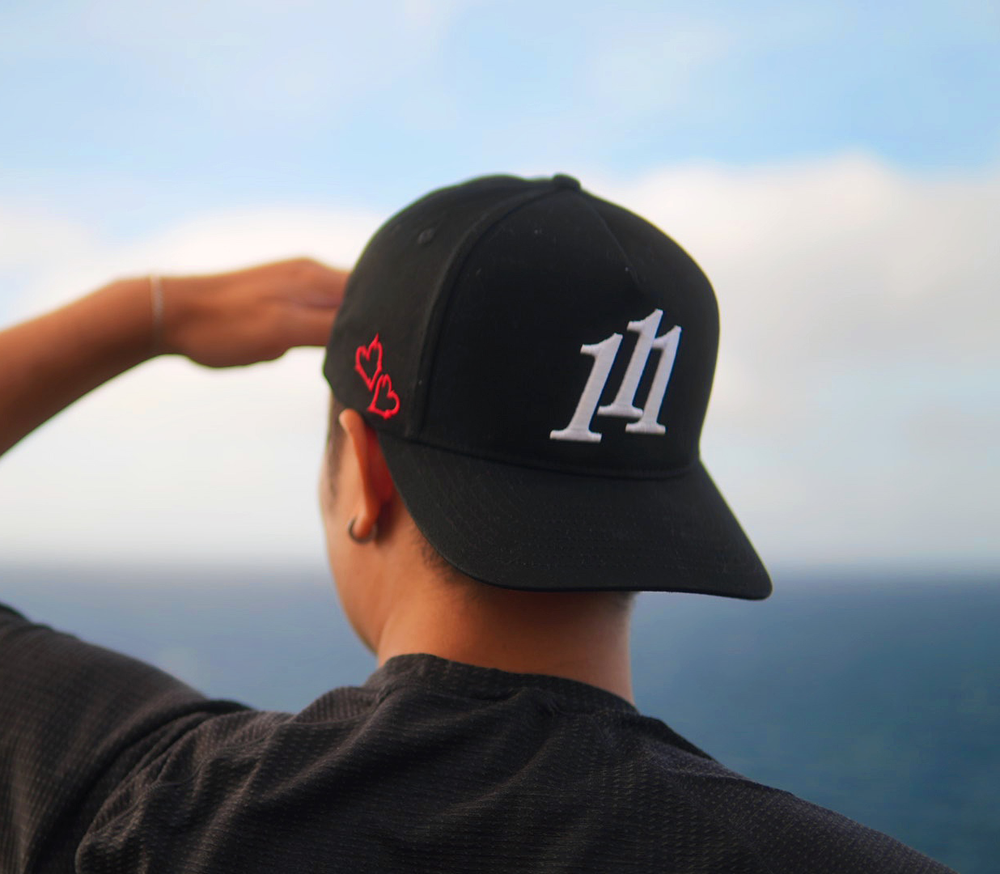 A person viewed from behind wearing a black baseball cap with a white stylized '111' angel number logo on the front and red heart embroidery on the side, with an outdoor ocean view in the background.