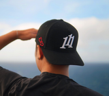 A person viewed from behind wearing a black baseball cap with a white stylized '111' angel number logo on the front and red heart embroidery on the side, with an outdoor ocean view in the background.
